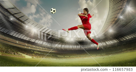 Female soccer player in red performs high jump kick on stadium field. Arena packed with spectators, and bright spotlights illuminate scene. Female soccer player in red performs high jump kick on stadium field. Arena packed with spectators, and bright spotlights illuminate scene. 116657983