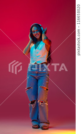 Full length photo of young beautiful woman dancing of joy with headphones in neon colors against magenta studio background, Trendy colors. Full length photo of young beautiful woman dancing of joy with headphones in neon colors against magenta studio background, Trendy colors. 116658020