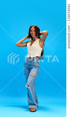 Cheerful young woman dressed casual attire with headphones on neck grimacing looking at camera against blue studio background. Trendy colors. 116658082