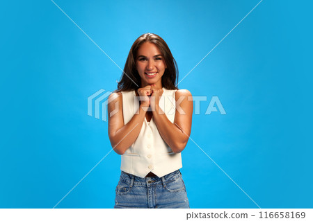 Portrait of young woman with long brown hair stands against bright blue background, exuding joy and excitement. Trendy colors. Portrait of young woman with long brown hair stands against bright blue background, exuding joy and excitement. Trendy colors. 116658169