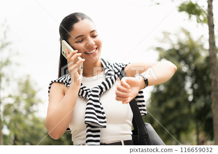 Chic young woman balancing student life and technology in the city, engrossed in her phone, symbolizing the blend of education and digital era. Checking time 116659032