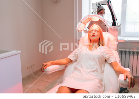 Relaxed blonde woman lying with closed eyes having LED light hair PDT treatment at luxury aesthetic clinic. Professional specialist adjusting innovation device for photodynamic therapy procedure 116659462