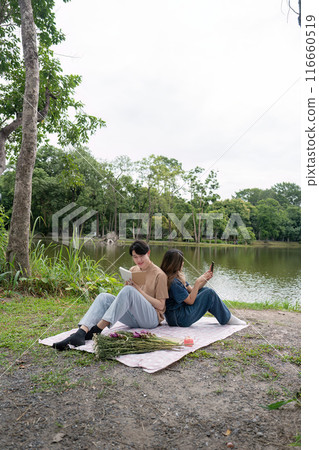 Young Couple Relaxing by a Serene Lake, Reading and Enjoying Nature on a Picnic Blanket in a Tranquil Park Setting Young Couple Relaxing by a Serene Lake, Reading and Enjoying Nature on a Picnic Blanket in a Tranquil Park Setting 116660519