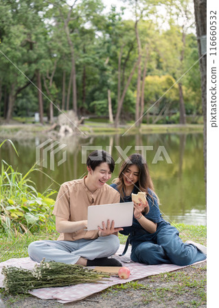 Happy Couple Enjoying a Picnic by the Lake with a Laptop and Snacks in a Serene Park Setting 116660532