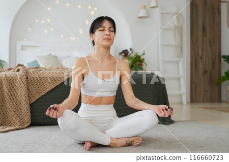 Yoga mindfulness meditation. Young healthy woman practicing yoga in living room at home. Woman sitting in lotus pose meditating smiling relaxing indoor. Girl doing breathing practice. Yoga at home 116660723