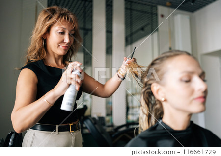 Lady client with long blonde hair getting stylish hairdo done at salon. Female hairdresser spraying hair setting liquid on blonde woman customer hair. Concept of professional fashion hairstyling. 116661729