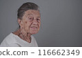Senior woman 90 years old smiles, laughs and shows a denture on a gray background. Elderly female with deep wrinkles and gray hair happy in a good mood studio portrait looking at camera 116662349