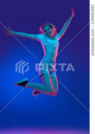 Young woman dressed in summer outfit jumps in mid-air of joy and happiness in neon light against gradient studio background. Trends. Young woman dressed in summer outfit jumps in mid-air of joy and happiness in neon light against gradient studio background. Trends. 116662882