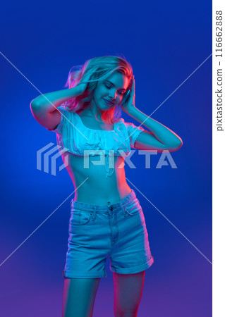 Young cheerful blonde woman radiates happiness while listening music in headphones in neon light against gradient studio background. Trends. 116662888