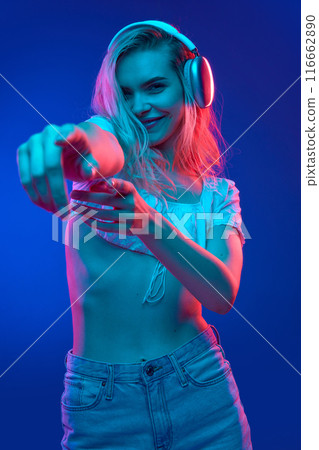 Young woman radiates happiness while listening music in headphones and singing in neon light against gradient studio background. Trends. Young woman radiates happiness while listening music in headphones and singing in neon light against gradient studio background. Trends. 116662890
