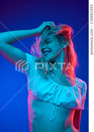 Joyful young woman posing with her hair playfully tossed, radiates happiness and carefree energy in neon light against gradient studio background. Trends. Joyful young woman posing with her hair playfully tossed, radiates happiness and carefree energy in neon light against gradient studio background. Trends. 116662893
