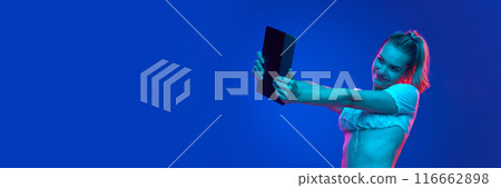 Banner. Young lady smiling to tablet screen in neon light against gradient studio background with negative space to insert text. Trends. Banner. Young lady smiling to tablet screen in neon light against gradient studio background with negative space to insert text. Trends. 116662898