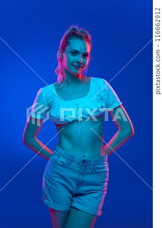 Confident young woman with slight smile, hands behind her back, and exudes sense of poise and charm in neon light against gradient studio background. Trends. Confident young woman with slight smile, hands behind her back, and exudes sense of poise and charm in neon light against gradient studio background. Trends. 116662912