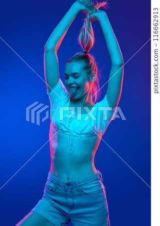 Young woman with raised arms as she playfully pulls her hair and sticks out her tongue in neon light against gradient studio background. Trends. Young woman with raised arms as she playfully pulls her hair and sticks out her tongue in neon light against gradient studio background. Trends. 116662913