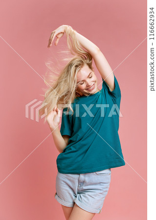 Joyful young woman with blonde hair, wearing t-shirt and shorts against pastel pink studio background. Her hair flows as she smiles, embodying happiness and freedom. Joyful young woman with blonde hair, wearing t-shirt and shorts against pastel pink studio background. Her hair flows as she smiles, embodying happiness and freedom. 116662934