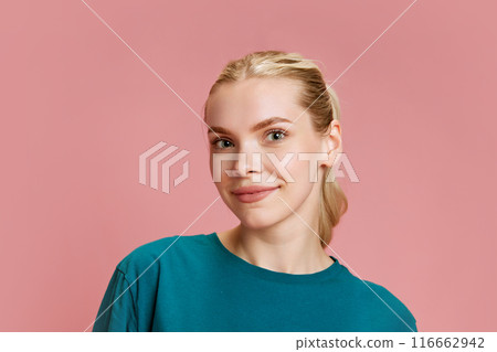 Young attractive blonde woman dressed blue T-Shirt looking at camera with light smile against pastel pink studio background. Young attractive blonde woman dressed blue T-Shirt looking at camera with light smile against pastel pink studio background. 116662942