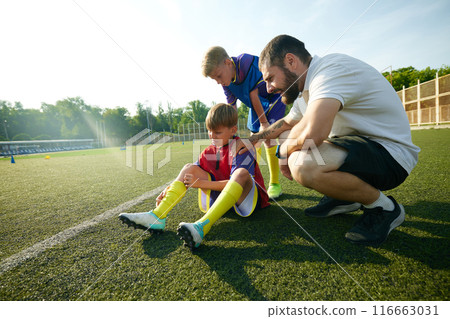 Coach providing assistance to child, boy sitting on field and feeling pain on legs, being injured during game. 116663031