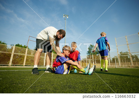 Coach providing assistance to child, boy sitting on field and feeling pain on legs, being injured during game. Coach providing assistance to child, boy sitting on field and feeling pain on legs, being injured during game. 116663032