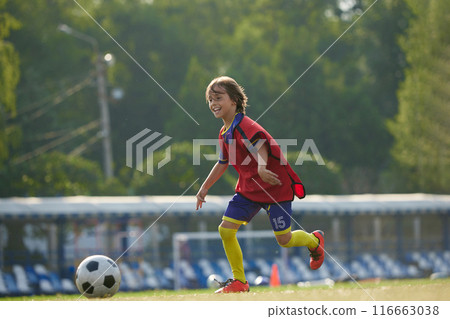 Concentrated boy, child in red and blue sportswear dribbling ball through field with focus, training soccer game. 116663038