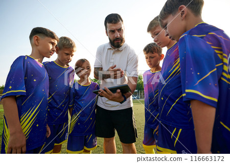 Coach discussing strategy with young soccer players, using a tablet to illustrate points. Outdoor game training Coach discussing strategy with young soccer players, using a tablet to illustrate points. Outdoor game training 116663172