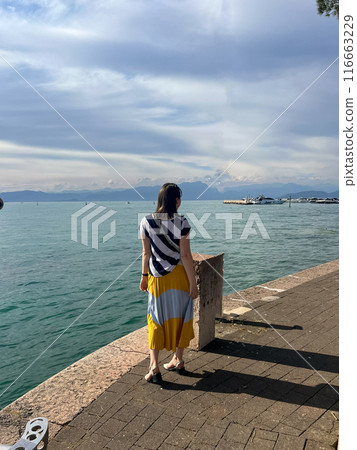 Woman in long dress enjoying stunning view over the Garda lake, seen from Peschiera del Garda Woman in long dress enjoying stunning view over the Garda lake, seen from Peschiera del Garda 116663229