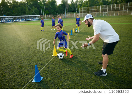 Boy practices dribbling through cones with focused encouragement from the coach. Outdoor stadium and evening game. Boy practices dribbling through cones with focused encouragement from the coach. Outdoor stadium and evening game. 116663263