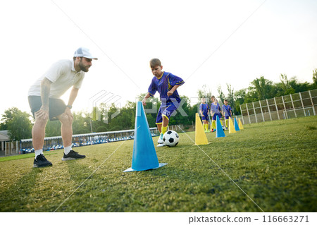 Young boy, child in motion, dribbling soccer ball around cones and receiving guidance from coach. Outdoor stadium game Young boy, child in motion, dribbling soccer ball around cones and receiving guidance from coach. Outdoor stadium game 116663271