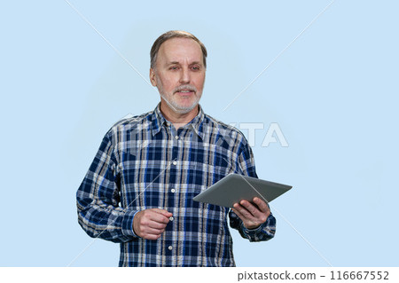 Portrait of a senior elder man holding tablet pc device. Casual blue checkered shirt. Isolated on pale blue background. Portrait of a senior elder man holding tablet pc device. Casual blue checkered shirt. Isolated on pale blue background. 116667552