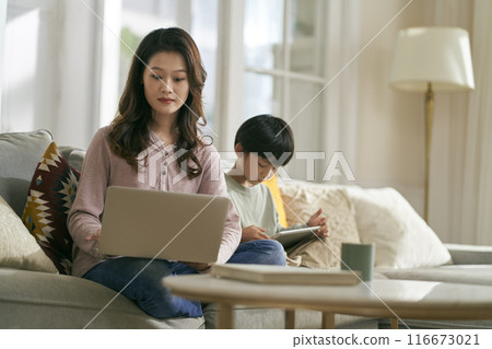 young asian mother working on laptop while son playing computer game using digital tablet at home young asian mother working on laptop while son playing computer game using digital tablet at home 116673021