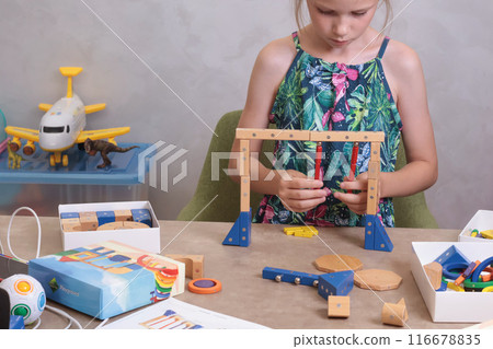 Girl assembles swing from magnetic wooden construction set. Toys and activiti children. Developmental activities at home. 116678835