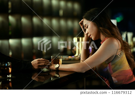 Young woman sitting at a dimly lit bar, lost in thought with a pensive expression and friend consoling her 116679312