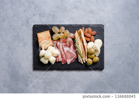 A plate with cheese, ham, mozzarella, olives, nuts, grapes and other snacks on a black stone board  116688845