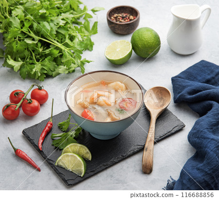 Tom yam soup with shrimp and chili pepper in a blue bowl on a light background with fresh vegetables 116688856