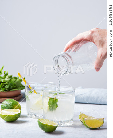a hand pours a lime mojito lemonade into a glass on the table with lime slices and fresh mint.  116688862