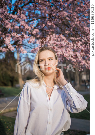 A woman poses under cherry blossoms in a white shirt during spring, creating a vibrant scene 116689139