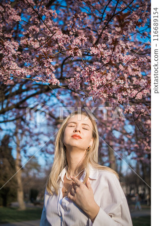 Fashion outdoor photo of beautiful woman with blond hair in elegant suit posing in spring flowering park with blooming cherry tree. Copy space and empty place for advertising text 116689154