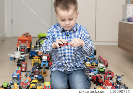 Concept of children's toys. A little boy, 4 years old, enthusiastically plays, sitting on the floor, with multi-colored small and large cars in the children's room. Concept of children's toys. A little boy, 4 years old, enthusiastically plays, sitting on the floor, with multi-colored small and large cars in the children's room. 116690725