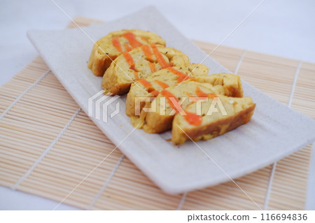 A white rectangular plate features neatly arranged slices of tamagoyaki, a Japanese rolled omelette. The omelette is drizzled with a bright orange sauce A white rectangular plate features neatly arranged slices of tamagoyaki, a Japanese rolled omelette. The omelette is drizzled with a bright orange sauce 116694836