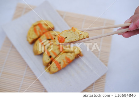 A white rectangular plate features neatly arranged slices of tamagoyaki, a Japanese rolled omelette. The omelette is drizzled with a bright orange sauce 116694840