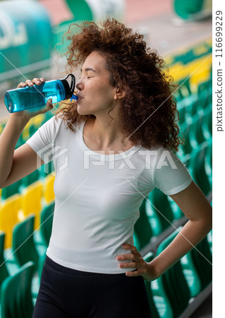 Beautiful curly girl in a white T-shirt and in black leggings drinks water at the stadium after a run 116699229