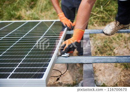 Close up of male workers in gloves installing solar panel on metal rails. Men hands mounting solar module for generating electricity through photovoltaic effect. Concept of renewable energy sources. 116700670