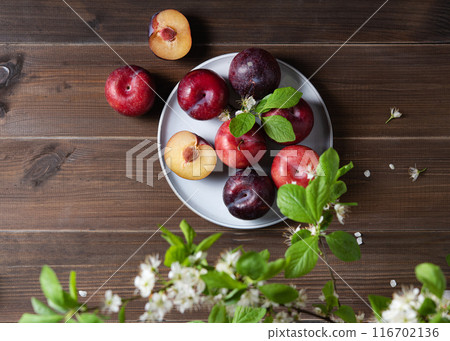 a few juicy red plums in blue plate with branch blossom flowers on a napkin on a wooden background.  116702136