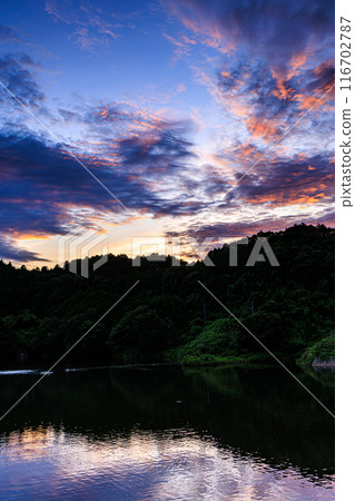 Evening scenery at Nunome Dam in the summer of 2024. A high-contrast sunset sky ranging from gray to golden madder appears above the dam lake. 116702787