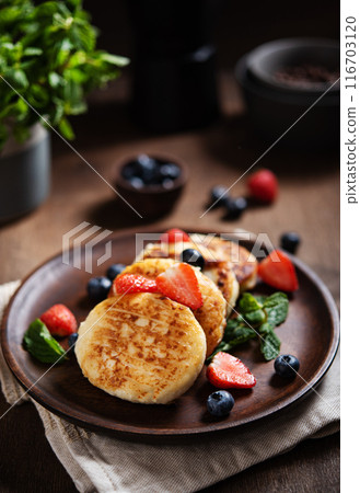 Cottage cheese pancakes or syrniki  with mint, strawberries and blueberries on a dark wooden table 116703120