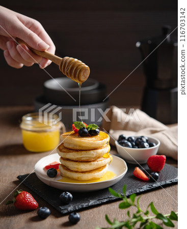 A hand pours honey on classic American pancakes in a stack with fresh berries 116703142