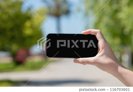 Hand holding smartphone against beautiful landscape in city park. Device screen with empty space 116703601