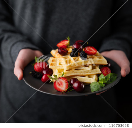 Hands holding a plate of Belgian waffles with fresh strawberries, blackberries, mint leaves on a dark background close up. 116703638