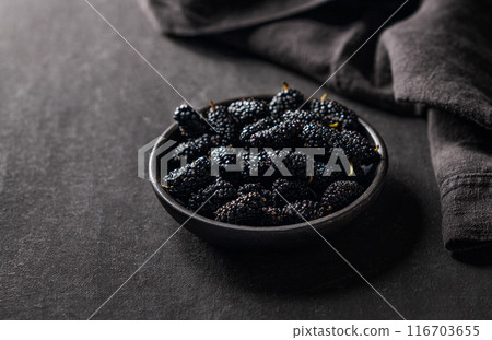 Fresh black mulberry with leaf in black wooden bowl on dark background with napkin. Fresh black mulberry with leaf in black wooden bowl on dark background with napkin. 116703655