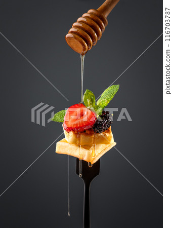 Wafer pieces with fresh strawberries, blackberries, mint leaves and dripping honey on a black fork Wafer pieces with fresh strawberries, blackberries, mint leaves and dripping honey on a black fork 116703787