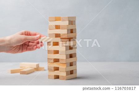A hand pulls out a piece from a wooden tower game on a light background. A hand pulls out a piece from a wooden tower game on a light background. 116703817